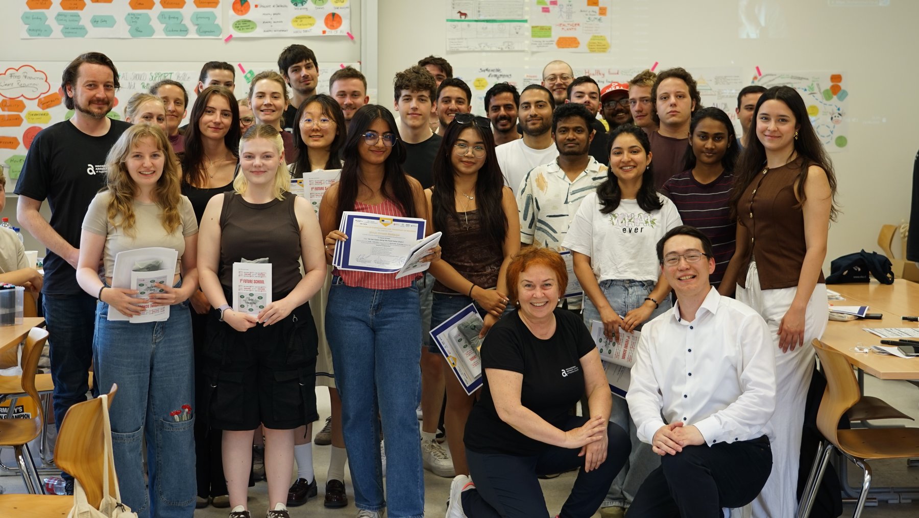 Gruppenfoto einer gemischten Gruppe von etwa 20 Personen im Klassenzimmer, die stolz Bücher und Urkunden halten. Zwei Personen sitzen vorne, der Rest steht hinter ihnen. Die Atmosphäre wirkt feierlich und erfolgreich.