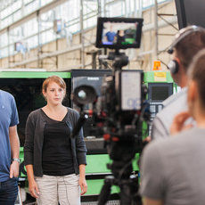 "Ein Filmteam auf einem Set. Zwei Personen stehen vor einer Kamera, während eine dritte Person hinter der Kamera steht und Anweisungen gibt. Im Hintergrund ist ein großer Raum mit Industrieelementen zu sehen."