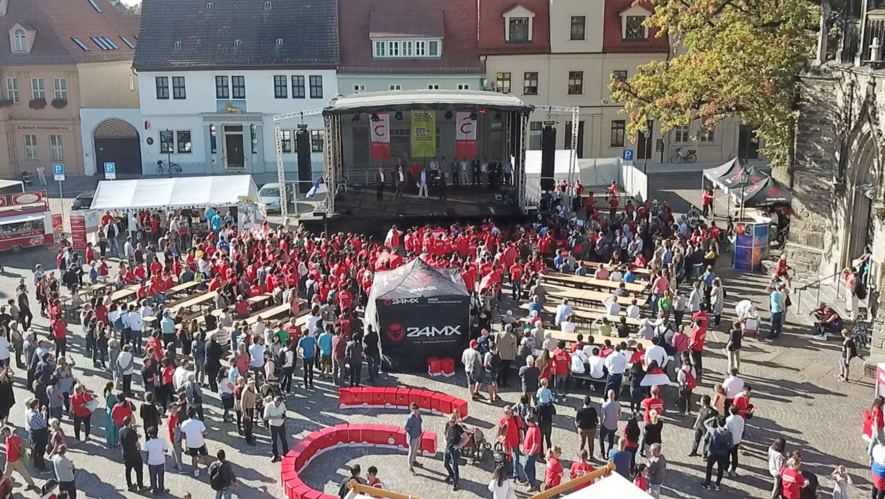 "Ein belebter Platz mit einer großen Menschenmenge, die sich um eine Bühne versammelt. Viele tragen rote Kleidung. Im Vordergrund steht ein schwarzes Fahrzeug mit der Aufschrift '24H MX'. Die Veranstaltung findet in einer historischen Stadt mit Gebäuden im Hintergrund statt."