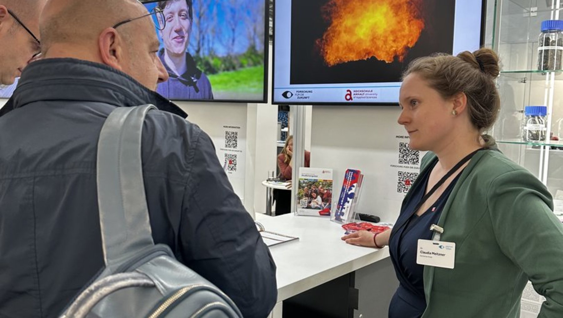 Zwei Personen auf einer Messe: Eine Mitarbeiterin in grünem Shirt erklärt an einem Infostand mit großem Bildschirm, der eine Explosion oder thermische Abbildung zeigt, während zwei Männer mit Rucksäcken interessiert zuhören.