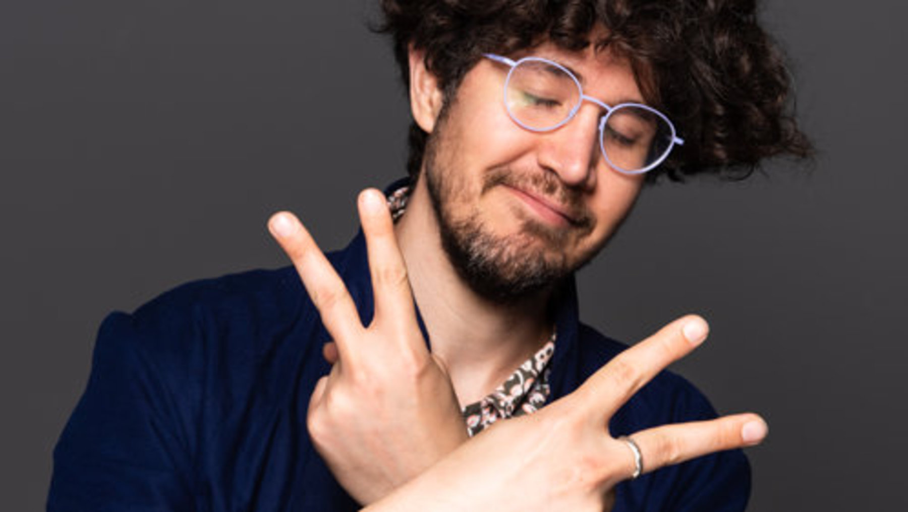 Ein lächelnder Mann mit kurzem, lockigem Haar und Brille zeigt mit beiden Händen die Peace-Zeichen-Gesten, während er ein blaues Jackett trägt.