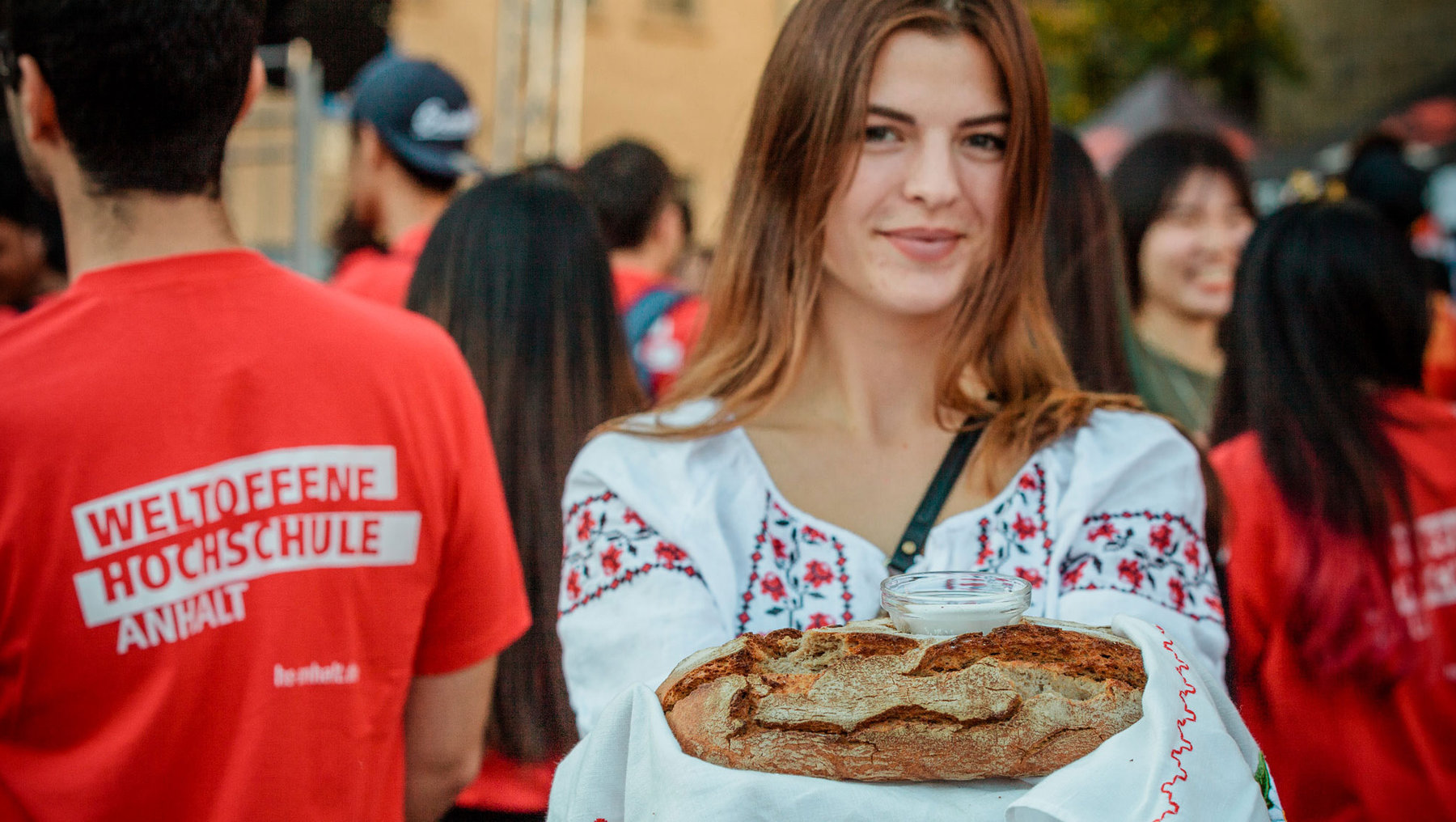 Eine Frau in traditioneller Tracht hält zwei Brotlaibe auf einem Tuch und daneben ein Glas, lächelnd bei einer Veranstaltung. Im Hintergrund tragen Teilnehmer rote T-Shirts mit der Aufschrift „Weltoffene Hochschule Anhalt“.