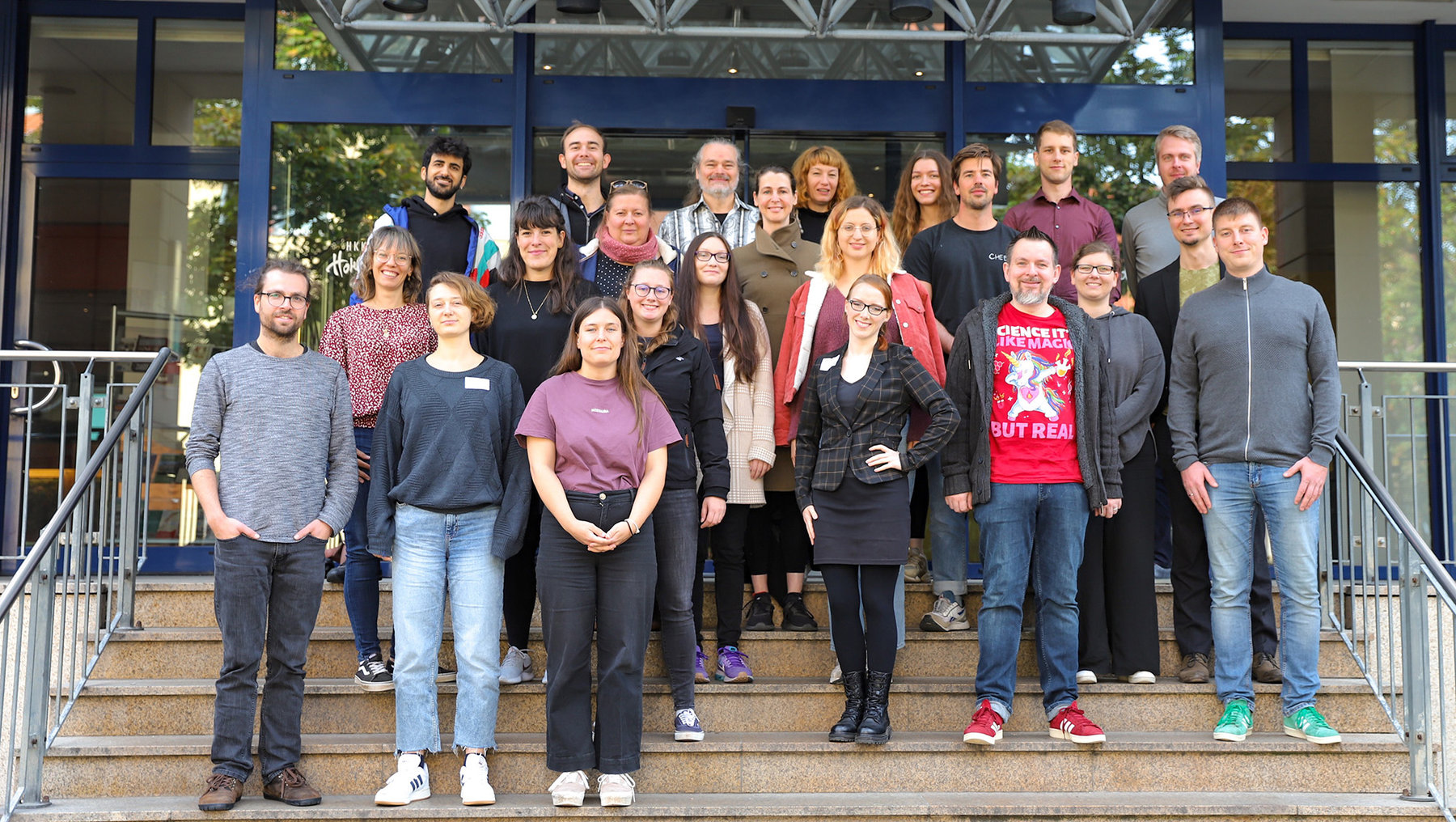 Gruppe von 20 Personen, die vor einem Gebäude mit Treppe und Geländer stehen, lächeln in die Kamera. Verschiedene Kleidungsstile, darunter Jacken, T-Shirts und ein T-Shirt mit Aufdruck.
