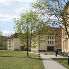 A large, multi-story building with a beige facade and brown roof, surrounded by a grassy area with several trees. A paved walkway leads towards the building. A person is walking along the path, and bicycles are parked near the building. The sky is partly cloudy.