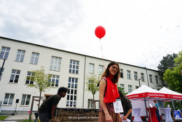 Eine Frau in rotem Oberteil hält einen roten Luftballon und lächelt fröhlich vor einem hellen Mehrfamiliengebäude mit vielen Fenstern. Im Hintergrund sind weitere Personen und ein Infostand mit Markise zu sehen.