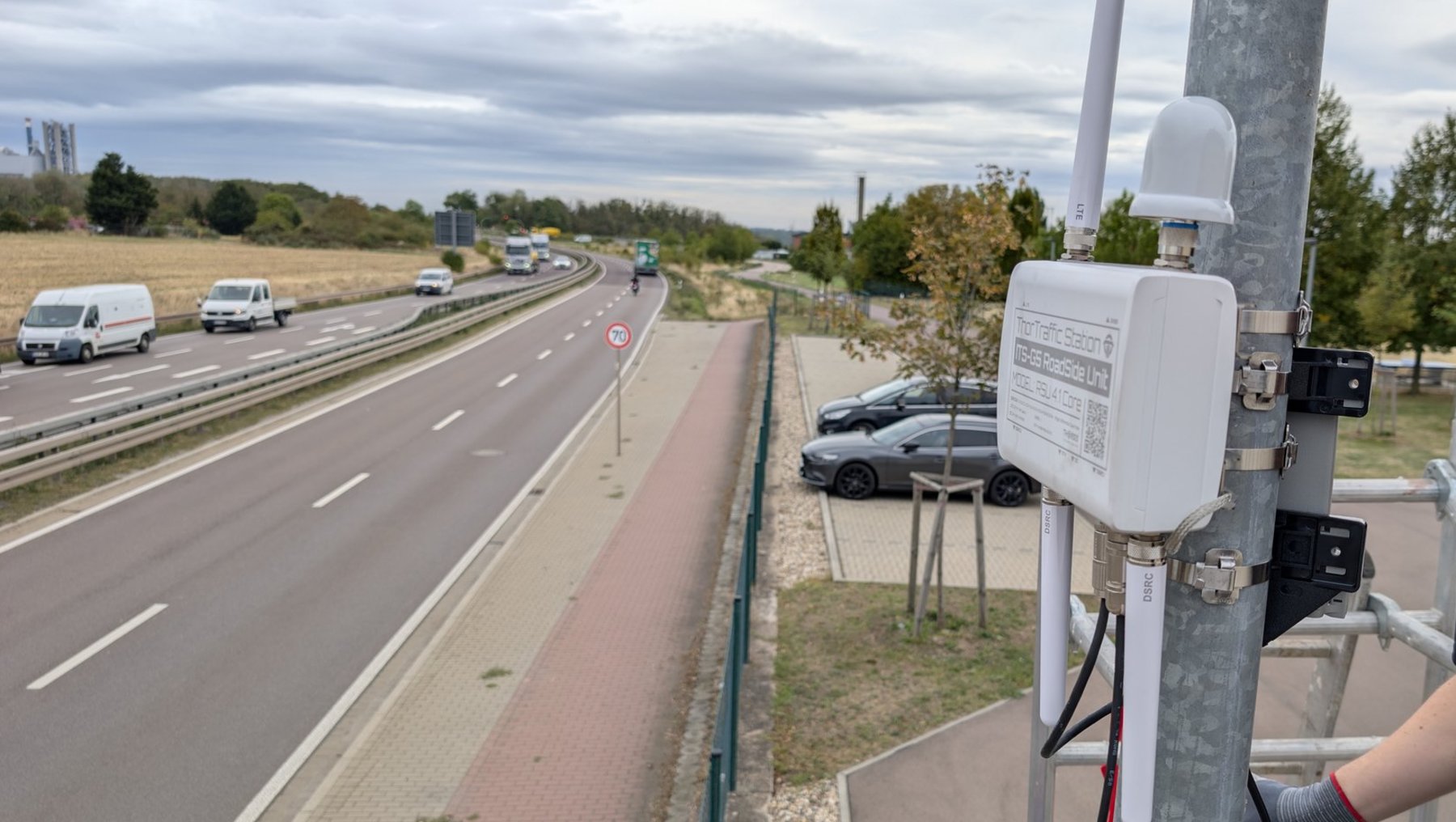 An der Straßenseite ist eine weiße Messvorrichtung an einem Pfahl befestigt, die mit Kabeln verbunden ist. Im Hintergrund verläuft eine mehrspurige Autobahn mit mehreren Fahrzeugen und einem Radweg.