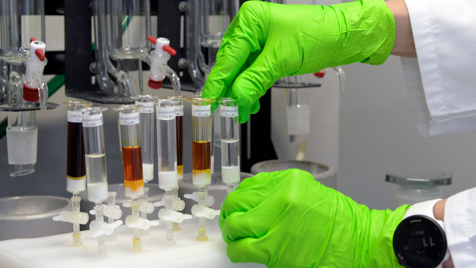 A person in a lab setting is handling multiple small test tubes containing different colored liquids, posing for an experiment or testing. Green gloves are worn for safety. Various lab equipment is present.