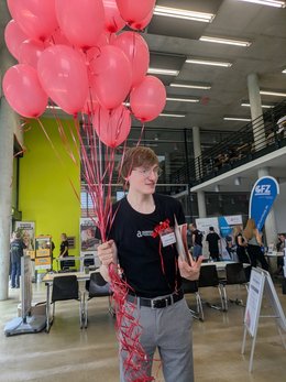 Ein junger Mann hält ein großes Bündel aus etwa 20 roten Luftballons, die mit einem Band zusammengebunden sind, und lächelt dabei. Er trägt ein schwarzes Shirt und eine Namensschleife.