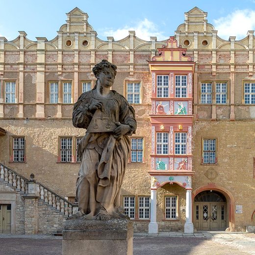 A statue of a robed figure holding a book stands in front of an ornate, historic building with multiple windows and decorative elements. The building features a staircase on the left side and colorful murals above the entrance. The architecture suggests a European style, possibly from the Renaissance or Baroque period.