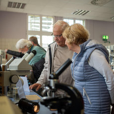 Zwei ältere Personen betrachten gemeinsam durch ein Mikroskop in einem hellen Laborraum. Im Hintergrund informieren sich weitere Seniorinnen und Senioren an verschiedenen Geräten und Stationen.