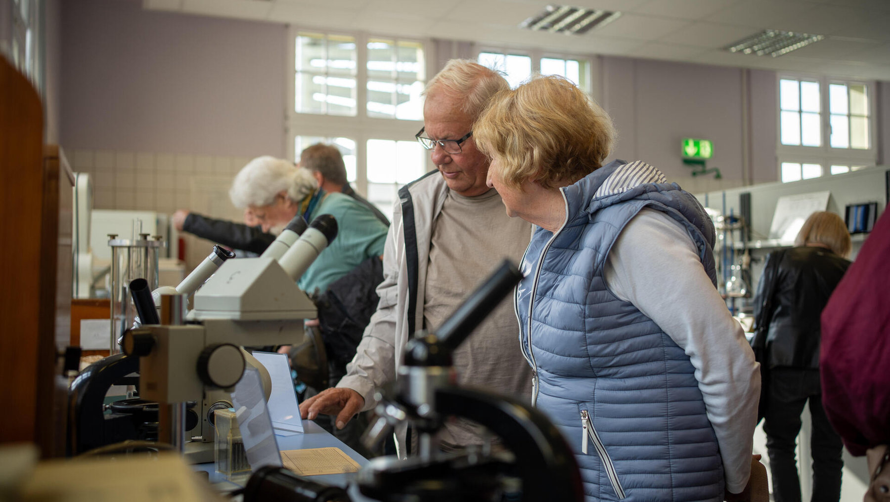 Zwei ältere Personen betrachten gemeinsam durch ein Mikroskop in einem hellen Laborraum. Im Hintergrund informieren sich weitere Seniorinnen und Senioren an verschiedenen Geräten und Stationen.