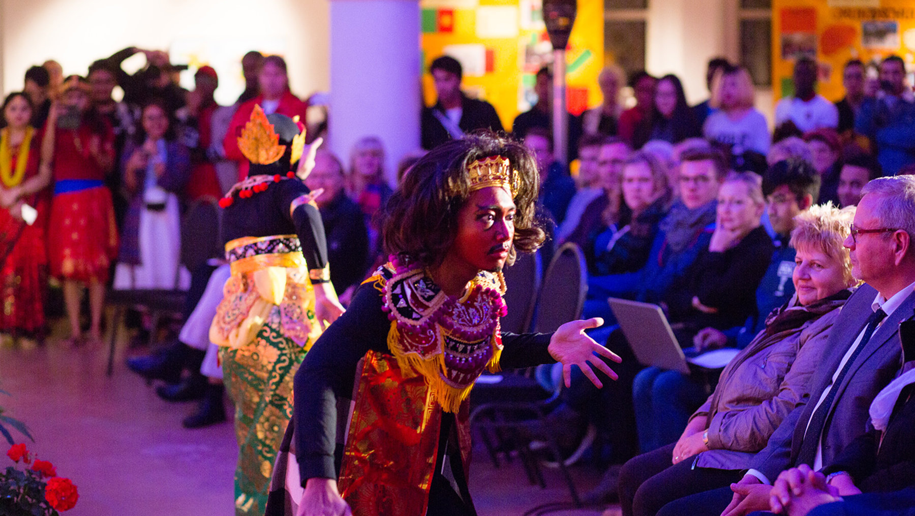 Eine bunte Theatervorstellung mit kostümierten Darstellern auf der Bühne. Die Schauspieler tragen farbenfrohe, traditionelle Gewänder und führen eine lebhafte Szene vor einem Publikum auf. Die Zuschauer sitzen auf Stühlen und verfolgen die Aufführung aufmerksam.
