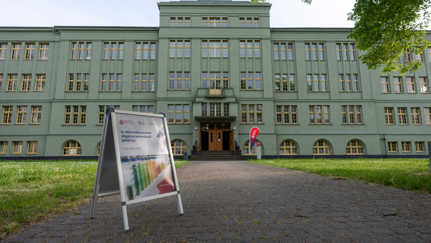 Plakat vor einem grünen Gebäude mit vielen Fenstern, das die Veranstaltung „4. Mitteldeutscher Algenstammtisch (MISTA)“ am 5. und 6. Mai 2026 ankündigt.