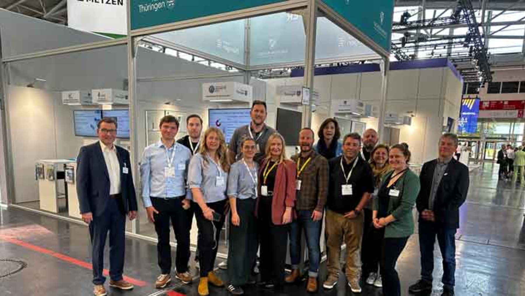 Gruppe von 14 Personen, darunter Männer und Frauen, steht lächelnd auf einer Messe vor einem Stand mit dem Namen „Metzen“ und „Thüringen“. Sie tragen Namensschilder und Business-Casual-Kleidung.