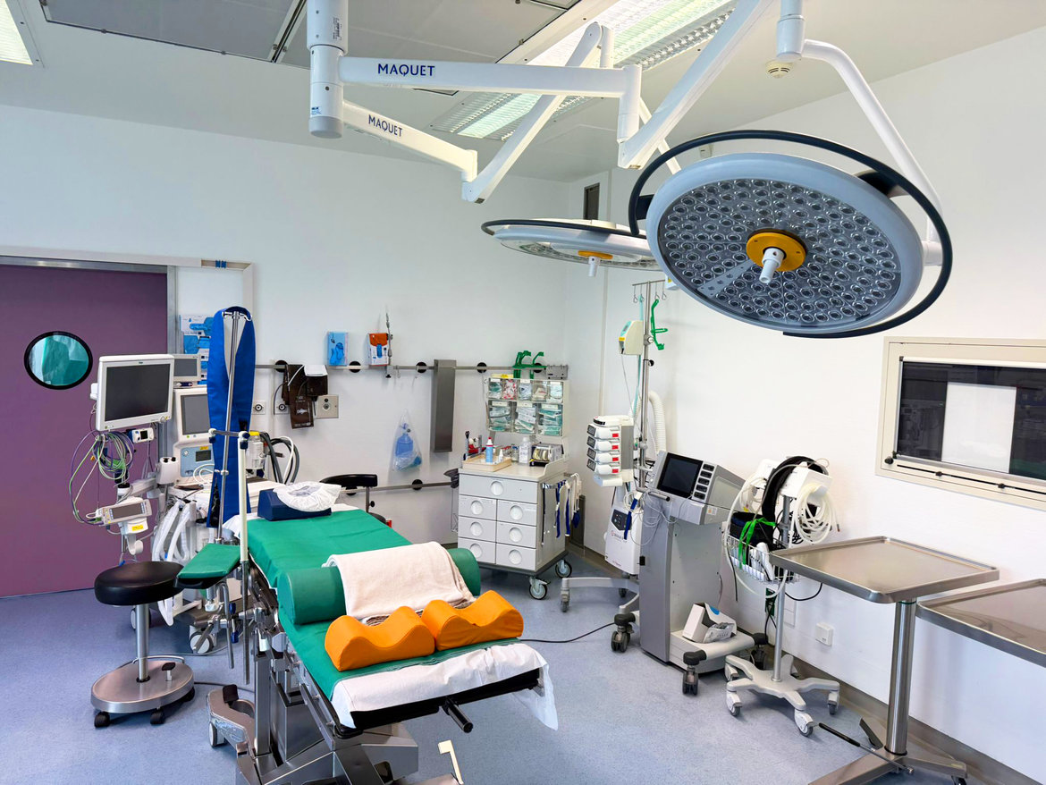 Moderner Operationssaal mit zentralem OP-Tisch, grünem OP-Tuch, zwei Operationsleuchten, medizinischen Geräten wie Monitoren und Infusionsständern sowie medizinischer Technik und Instrumenten an den Wänden.