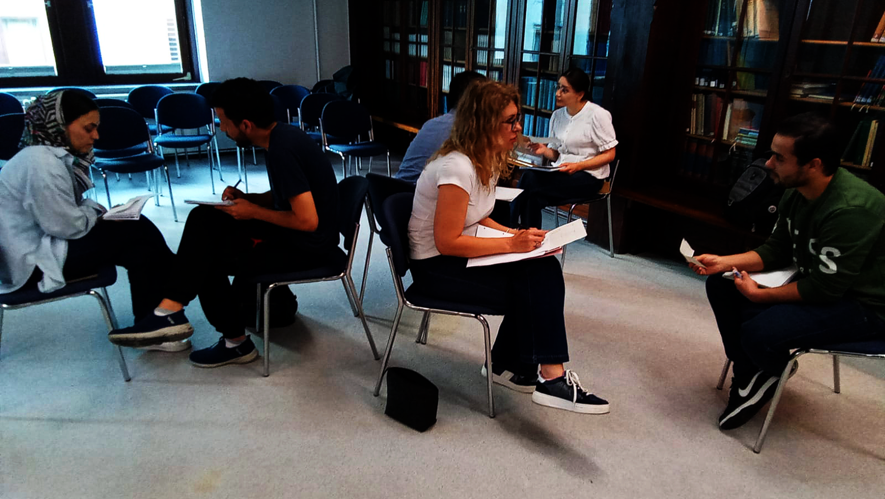 Fünf Personen sitzen in einem Kreis auf Stühlen in einem hellen Raum, vermutlich einer Bibliothek. Sie scheinen sich in einer Besprechung oder Diskussion zu befinden, wobei alle Notizen machen oder Papiere halten.