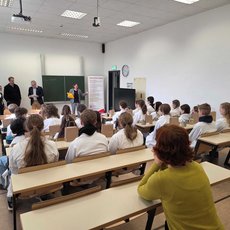 Ein Vortrag oder eine Präsentation findet in einem hellen Hörsaal statt. Vier Personen stehen vorne an der Bühne, während etwa 20 Schüler auf Bänken aufmerksam zuhören. Im Hintergrund sind eine Tafel und ein Plakat mit Informationen zu sehen.