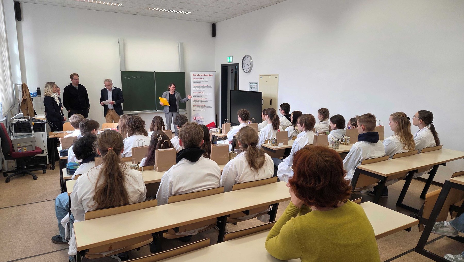 Ein Vortrag oder eine Präsentation findet in einem hellen Hörsaal statt. Vier Personen stehen vorne an der Bühne, während etwa 20 Schüler auf Bänken aufmerksam zuhören. Im Hintergrund sind eine Tafel und ein Plakat mit Informationen zu sehen.