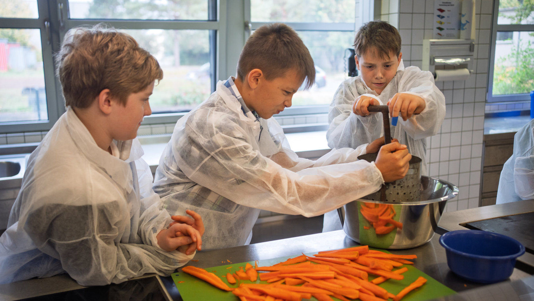 Drei Kinder in Kochjacken schneiden und verarbeiten Karotten in einer Küchenumgebung. Ein Kind rührt in einem Topf, während zwei andere Karotten schneiden und zerkleinern.