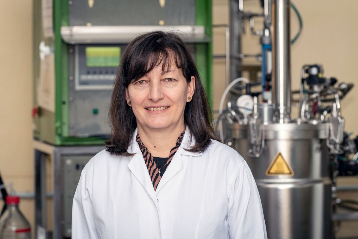 Eine lächelnde Person mit kurzem, dunklem Haar und weißem Laborkittel steht vor wissenschaftlicher Laborausrüstung, darunter ein Gerät mit Bedienfeld und ein silbriger Behälter mit Warnsymbol.