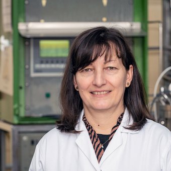 Eine lächelnde Person mit kurzem, dunklem Haar und weißem Laborkittel steht vor wissenschaftlicher Laborausrüstung, darunter ein Gerät mit Bedienfeld und ein silbriger Behälter mit Warnsymbol.