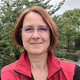 Eine Frau mit kurzen roten Haaren und einer Brille lächelt in die Kamera. Sie trägt eine rote Jacke mit schwarzem Reißverschluss. Im Hintergrund sind Bäume und ein bewölkter Himmel zu sehen.