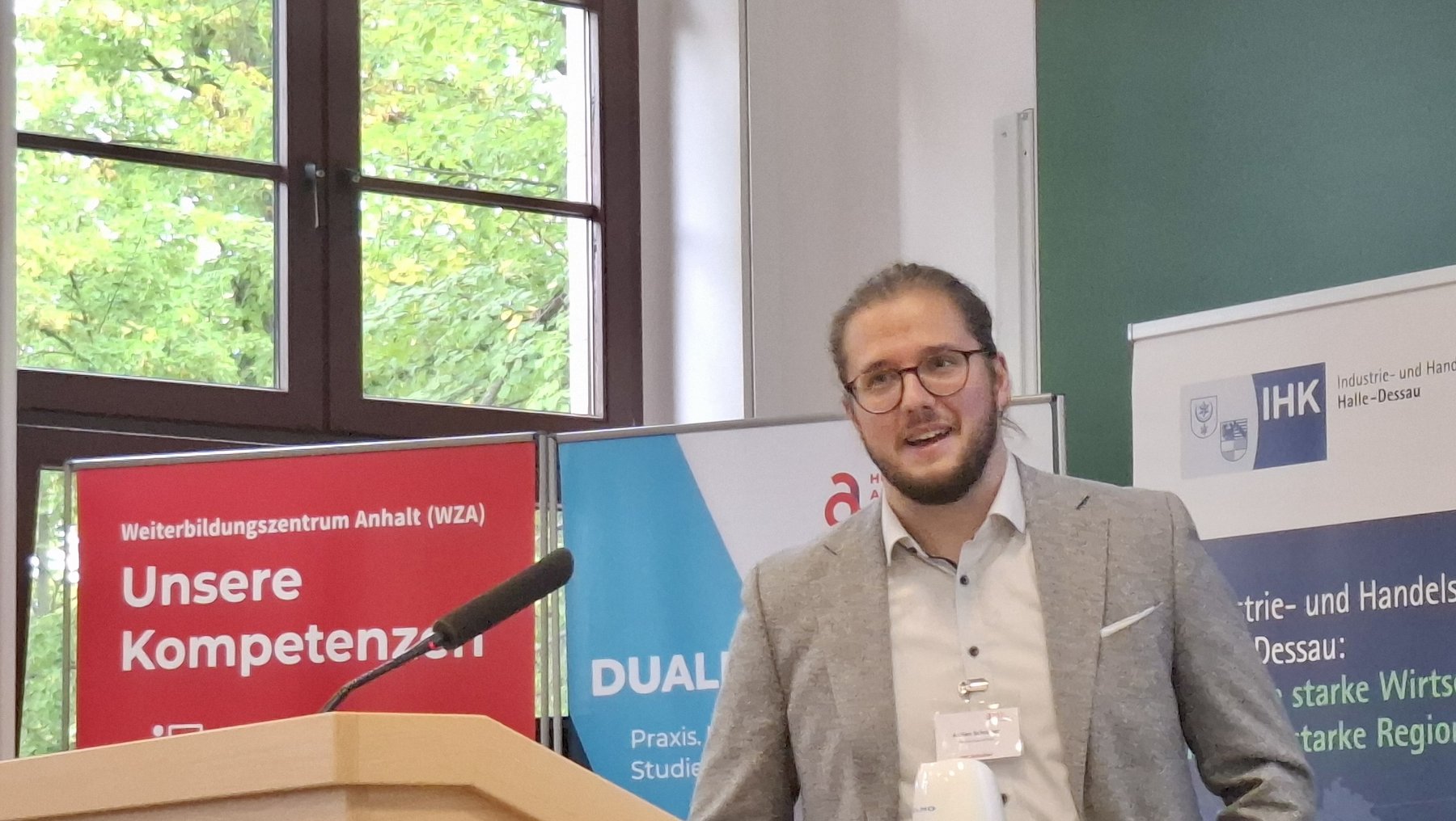 Ein Mann steht hinter einem Rednerpult und hält eine Rede. Er trägt eine Jacke und ein Namensschild. Auf dem Pult liegt ein Buch mit dem Titel „Unsere Kompetenzen“ sowie ein Mikrofon. Im Hintergrund sind Plakate mit Texten zu dualer Ausbildung und Wirtschaftsförderung zu sehen.