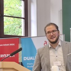 Ein Mann steht hinter einem Rednerpult und hält eine Rede. Er trägt eine Jacke und ein Namensschild. Auf dem Pult liegt ein Buch mit dem Titel „Unsere Kompetenzen“ sowie ein Mikrofon. Im Hintergrund sind Plakate mit Texten zu dualer Ausbildung und Wirtschaftsförderung zu sehen.