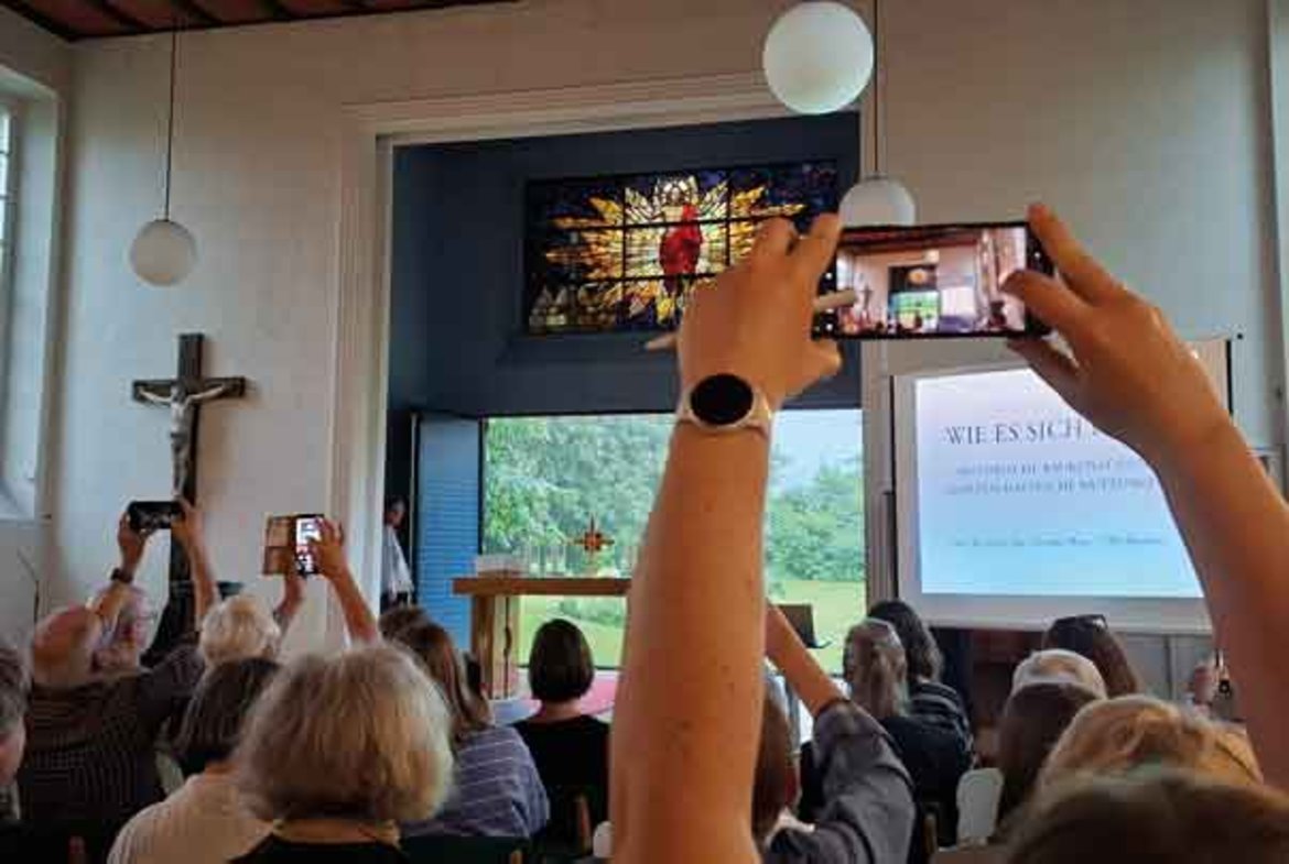 Menschen in einem Raum mit buntem Kirchenfenster im Hintergrund fotografieren mit Handys eine Präsentation auf Leinwand, auf der der Titel „Wie es sich gehört“ zu sehen ist. Mehrere Personen halten ihre Arme nach oben.