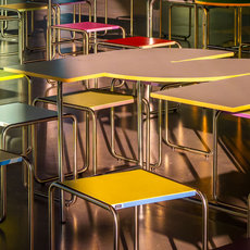 Mehrere bunte Tische und Stühle in einer Schul- oder Mensa-Kantine, farblich abgestimmt in Gelb, Blau, Grün, Rot und Grau, angeordnet in Reihen.