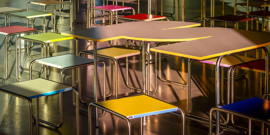 Mehrere bunte Tische und Stühle in einer Schul- oder Mensa-Kantine, farblich abgestimmt in Gelb, Blau, Grün, Rot und Grau, angeordnet in Reihen.
