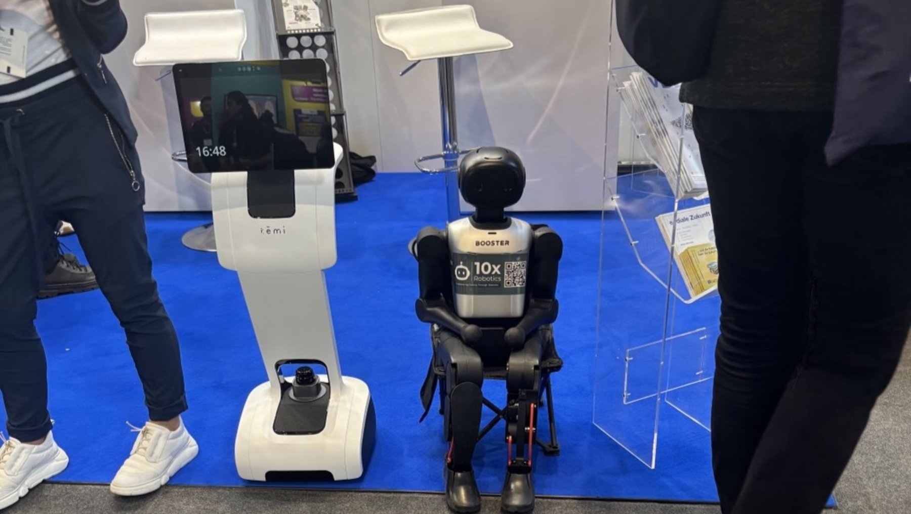 Zwei Roboter auf einer Messe: Ein humanoider Roboter mit Rollstuhl und schwarzer Kleidung, der „10x“ auf der Brust trägt, steht neben einem größeren, weißen Roboter mit Bildschirm und Bedienelementen.