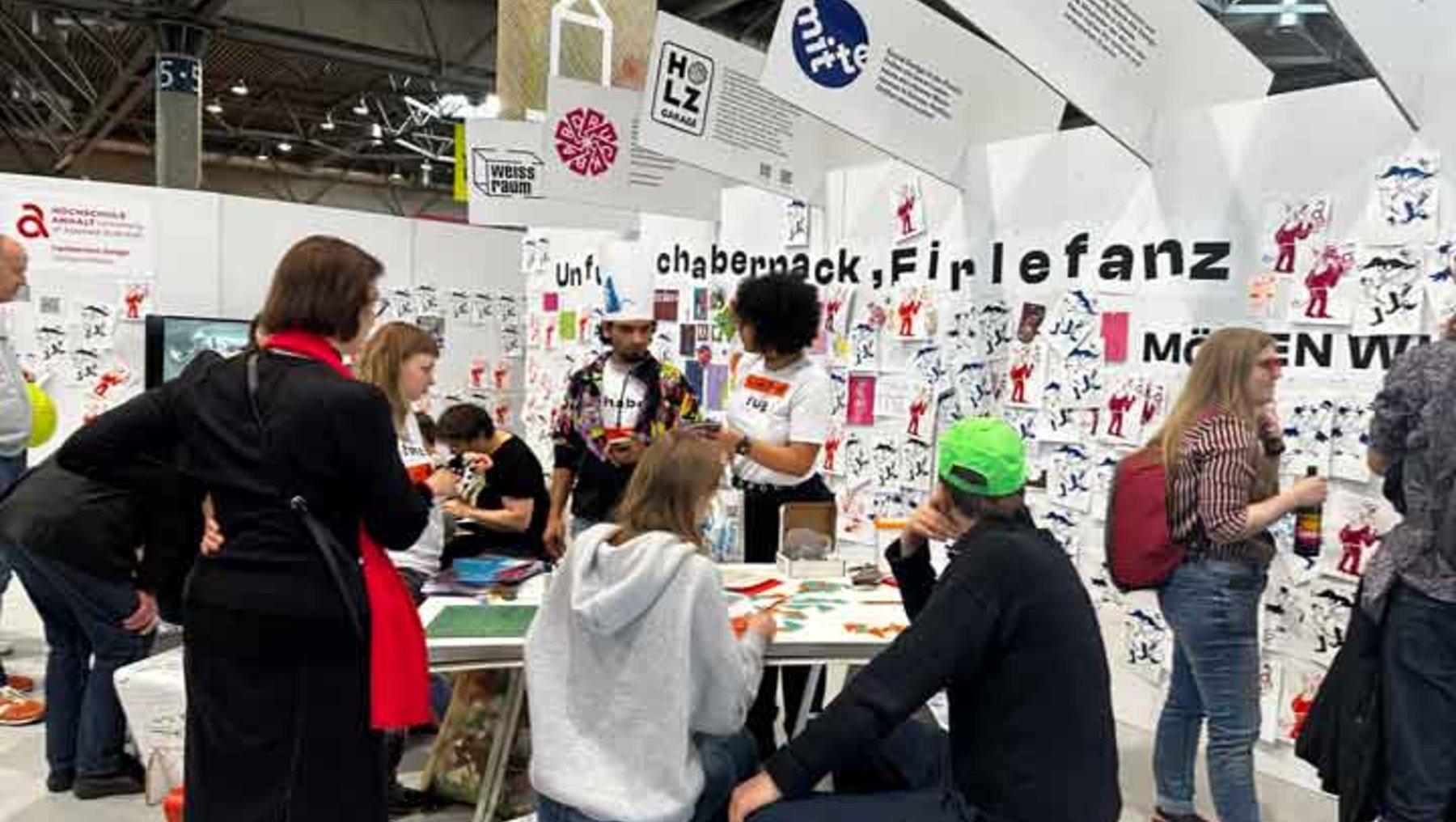 Menschen auf einer Messe besuchen einen bunten Infostand mit Postern und Plakaten zu verschiedenen Themen wie „chabempack“, „Firlefanz“ und „Möwen VW“. Einige sitzen an Tischen, andere stehen und unterhalten sich, während im Hintergrund weitere Besucher zu sehen sind.