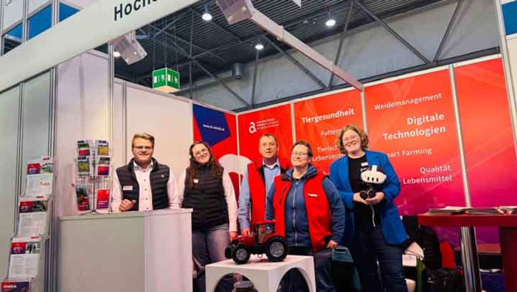 Vier Personen stehen lächelnd vor einem Messestand mit dem Thema digitale Technologien in der Landwirtschaft, insbesondere Tiergesundheit und Qualitätsmanagement. Im Vordergrund ist ein kleiner Traktor mit einem Roboterarm zu sehen.