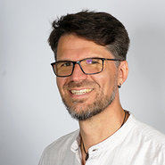Lächelnder Mann mit Brille, kurzem dunklem Haar und leichtem Bart, der ein helles Hemd trägt.