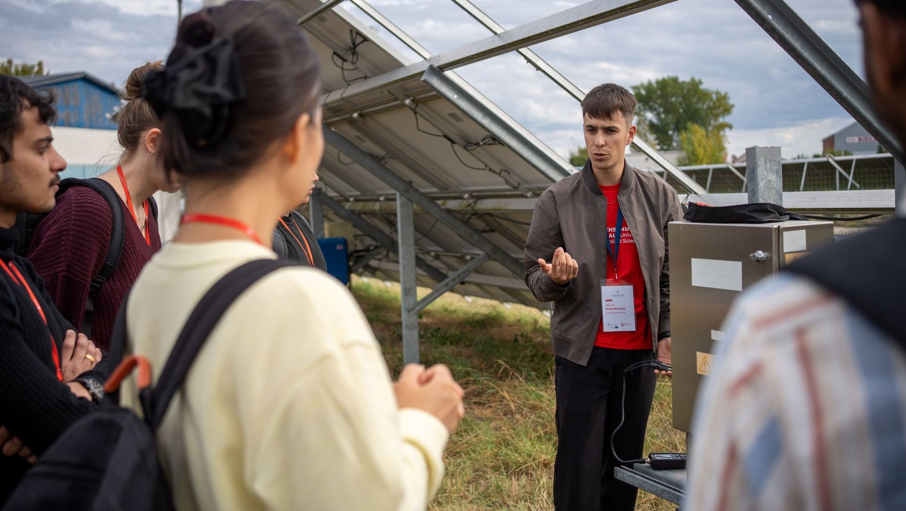 Eine Gruppe von Menschen steht vor einer großen Solaranlage auf einem Feld. Ein Mann im roten Shirt erklärt mit Gesten etwas über die Solarmodule, während die anderen aufmerksam zuhören.