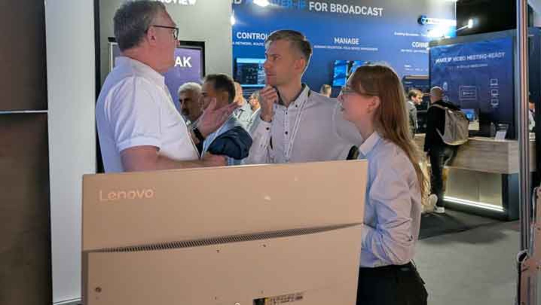 Drei Personen stehen vor einem Lenovo-Produktstand auf einer Messe. Ein Mann mit weißem Hemd und zwei weitere Personen, ein Mann und eine Frau, unterhalten sich. Im Hintergrund sind weitere Messebesucher und Infoschilder zu sehen.