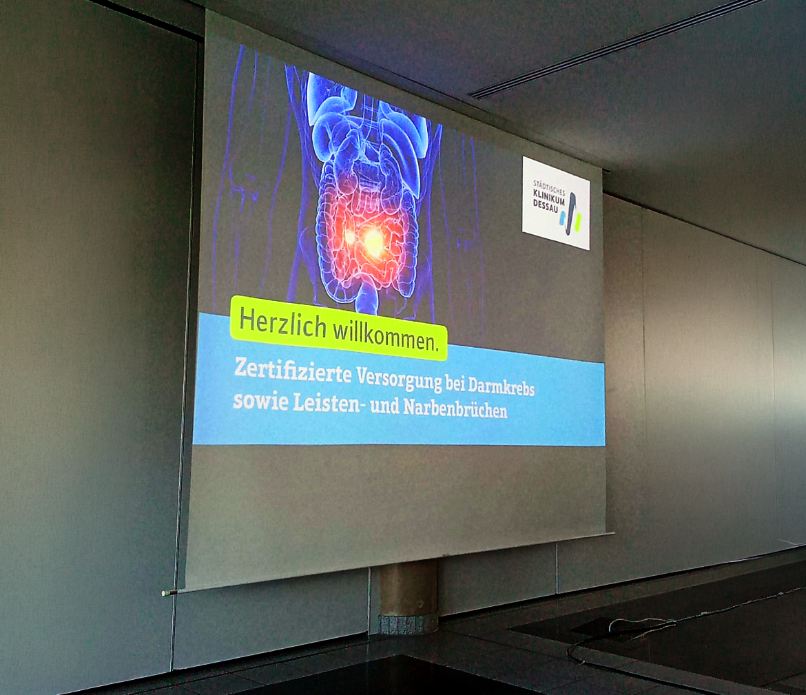 Willkommensbildschirm mit medizinischem Fokus: Herzlich willkommen zur zertifizierten Versorgung bei Darmkrebs sowie Leisten- und Narbenbrüchen, begleitet von einer schematischen Darstellung des menschlichen Unterleibs mit hervorgehobenem Darmbereich.