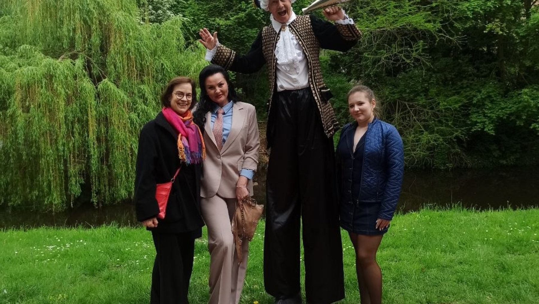 Vier Personen stehen im Freien und lächeln in die Kamera: eine Person in historischer Kleidung mit Dreispitz und Trompete, zwei Frauen in eleganter Kleidung und eine junge Frau in moderner Jacke.