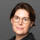 Eine Frau mit kurzen, dunklen Haaren und runden Brillengläsern trägt ein schwarzes Oberteil. Sie blickt leicht zur Seite und lächelt. Der Hintergrund ist grau und unauffällig.