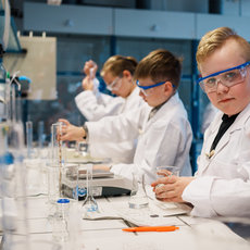 Drei Jugendliche in weißer Laborkleidung und Schutzbrillen führen im Chemielabor ein Experiment durch. Sie halten Reagenzgläser und arbeiten konzentriert an einem Labortisch mit verschiedenen Glasgeräten und Flüssigkeiten.