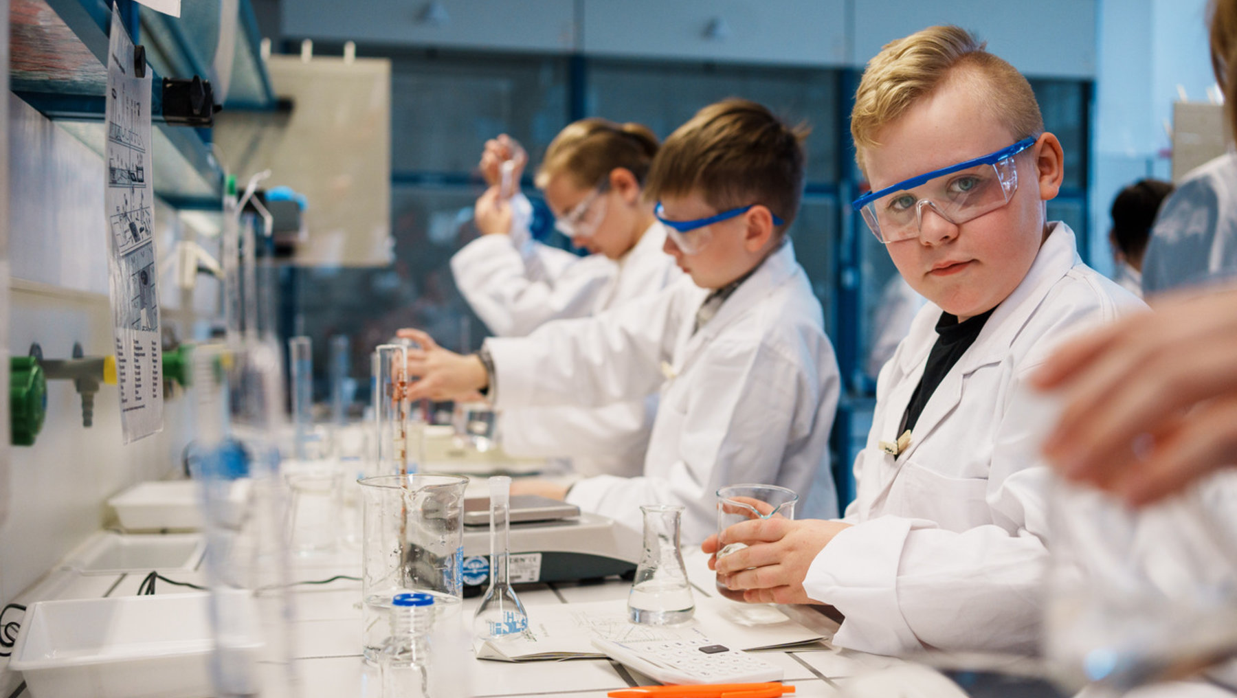 Drei Jugendliche in weißer Laborkleidung und Schutzbrillen führen im Chemielabor ein Experiment durch. Sie halten Reagenzgläser und arbeiten konzentriert an einem Labortisch mit verschiedenen Glasgeräten und Flüssigkeiten.