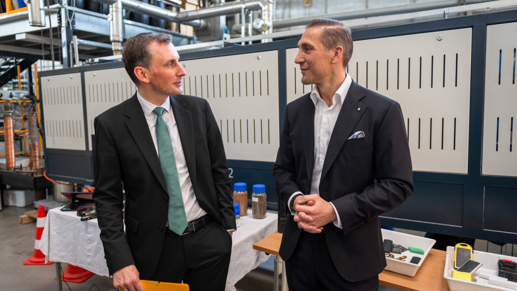 Zwei Herren in Anzügen unterhalten sich in einer industriellen Produktionshalle mit Maschinen im Hintergrund, während einer einen Ordner hält und der andere eine Uhr trägt.