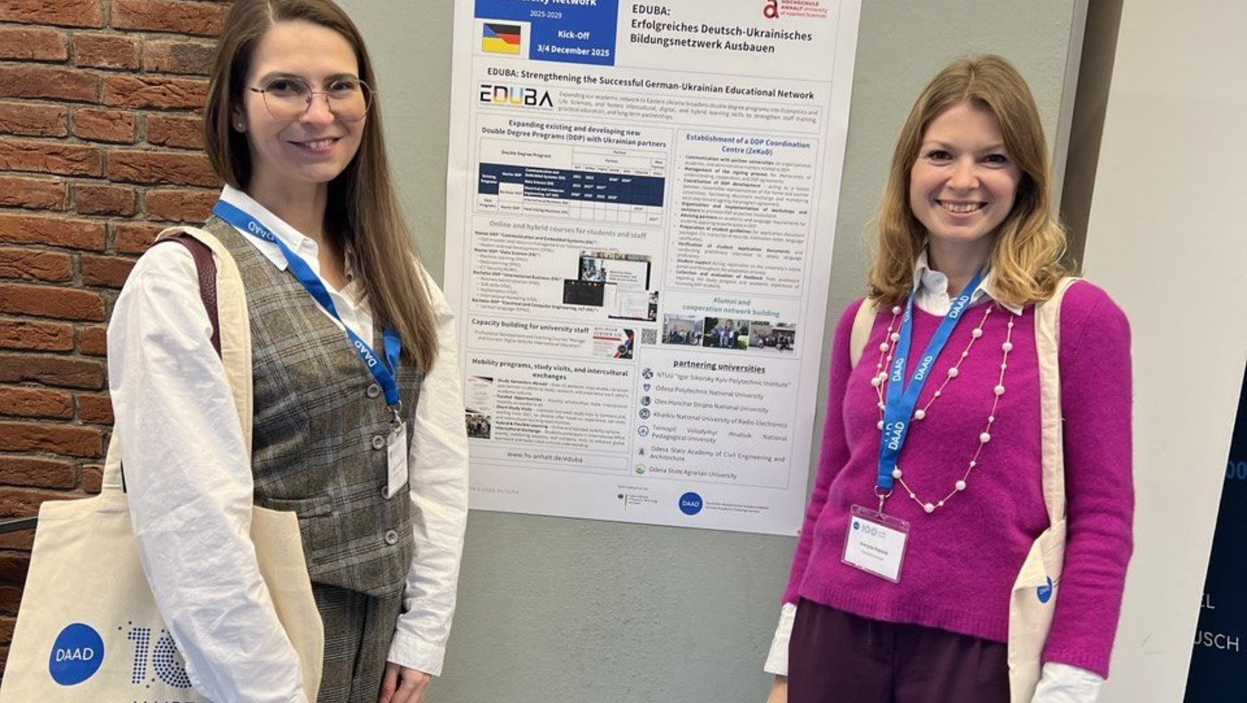Zwei Frauen stehen vor einem Poster an einer Wand. Beide tragen Namensschilder und Lächeln in die Kamera. Die linke Frau trägt einen gestreiften Overall und eine blaue Lanyard, während die rechte Frau einen pinken Pullover und eine violette Hose trägt. Sie präsentieren möglicherweise ihre Forschungsergebnisse.