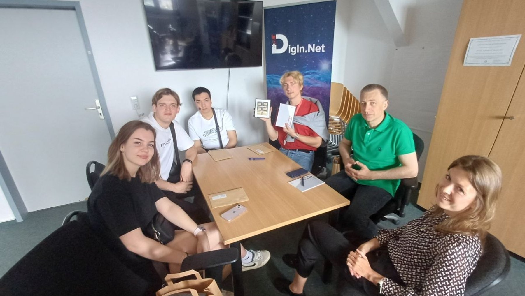Fünf Personen sitzen in einem Besprechungsraum um einen Tisch, lächeln in die Kamera und wirken entspannt. Im Hintergrund ist ein Banner mit dem Logo „Digin.Net“ zu sehen.
