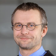 Porträt eines lächelnden Mannes mit kurz geschnittenen braunen Haaren, rundem Gesicht, Brille mit dünnen Metallgestellen und blauem Hemd.
