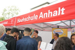 Ein Stand der Hochschule Anhalt mit rotem Zelt, auf dem „Hochschule Anhalt“ und Standorte wie Bernburg, Dessau, Köthen geschrieben sind. Mehrere Personen stehen im Gespräch, darunter eine Person mit Informationsmaterial in der Hand.