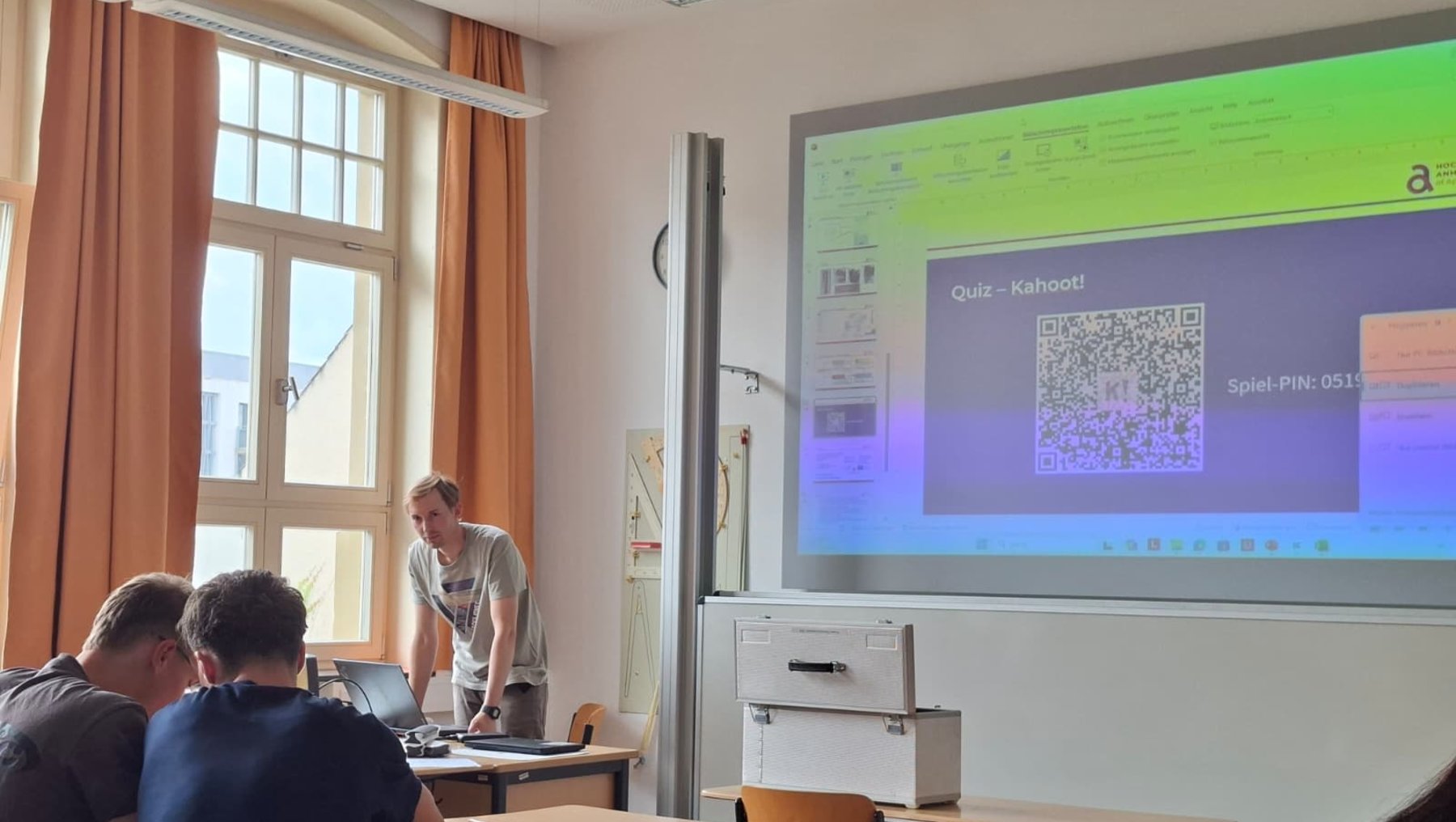 Ein Klassenzimmer mit drei Schülern, die an Tischen mit Laptops sitzen. Ein Lehrer steht vor einem großen Bildschirm, auf dem ein Quiz mit dem Titel Kahoot! und ein QR-Code für das Spiel angezeigt wird.
