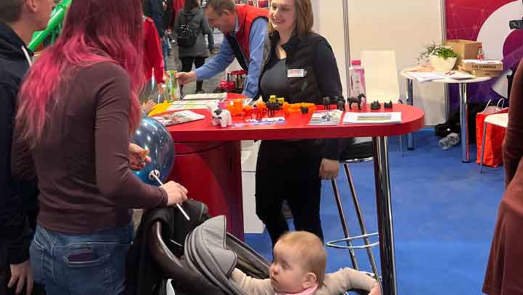 Eine Frau mit einem Baby im Tragegeschirr steht an einem Infostand. Die Frau hinter dem Tisch hält ein buntes, aufgeblasenes Spielzeug und bietet vermutlich eine Aktivität oder Mitmachstation für Kinder an.