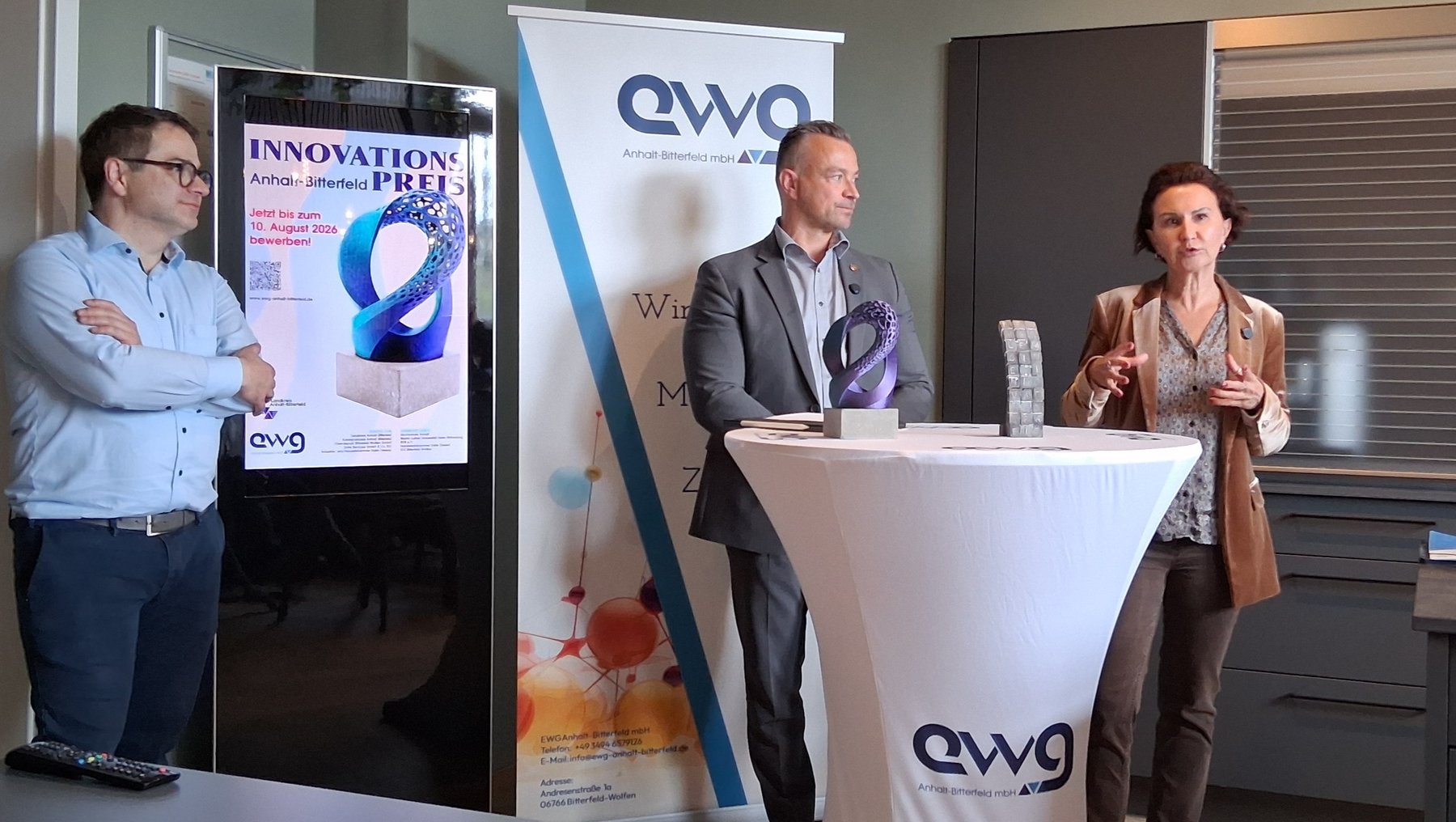 Drei Personen auf einer Bühne: Ein Mann mit verschränkten Armen, ein Mann in Anzug, der hinter einem Podium mit Mikrofon spricht, und eine Frau in Businesskleidung, die ebenfalls spricht. Im Hintergrund ein Banner mit „Innovationspreis“ und dem Logo „ewg“.